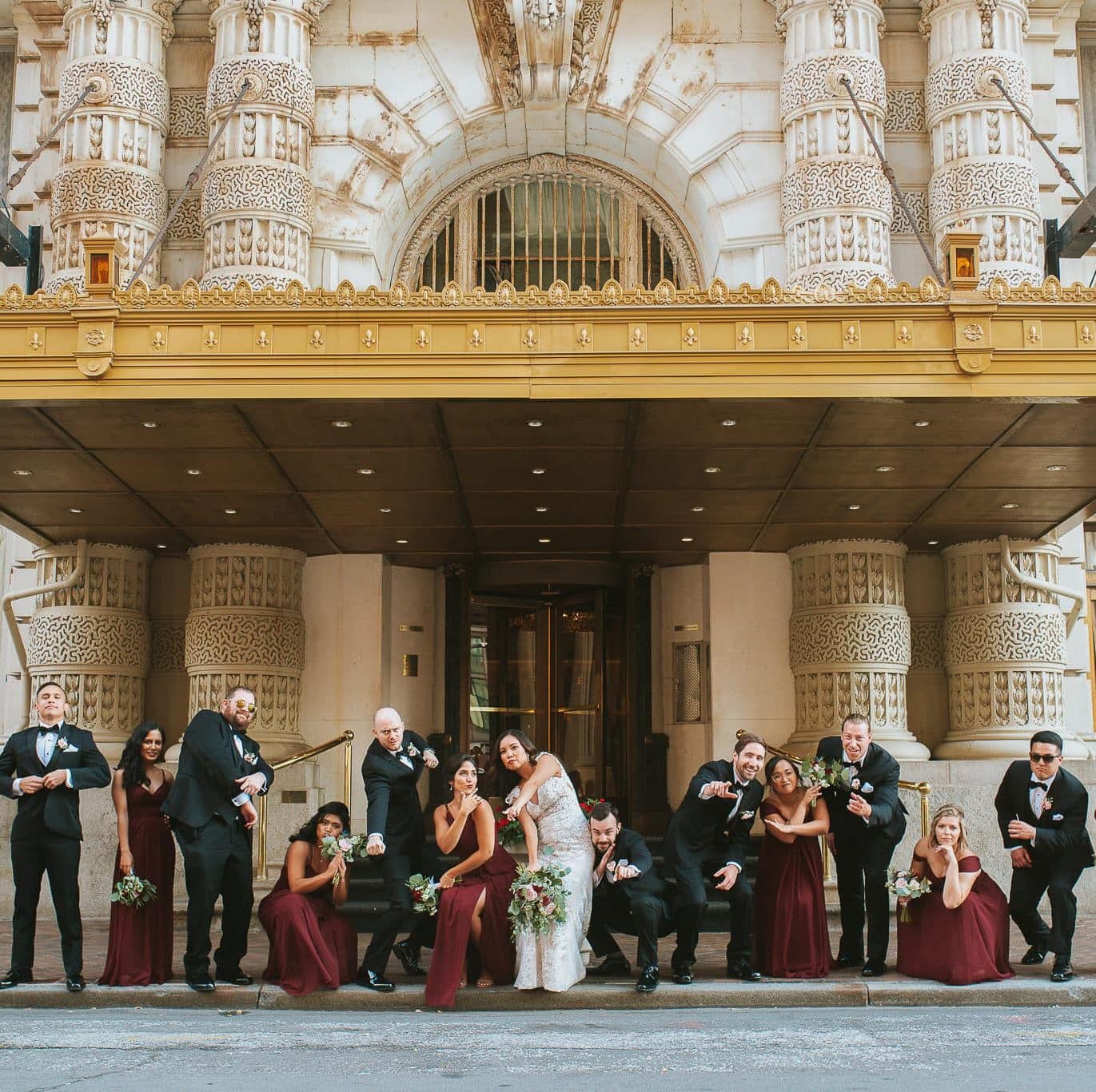 Wedding party posing playfully on the steps beneath the ornate gold marquee of the Belvedere Hotel in Baltimore